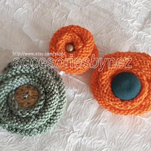 May include: Three handmade knitted fabric flower brooches. One is green with a brown button center, and two are orange with a brown button center and a teal button center.