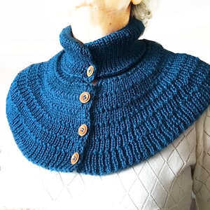 May include: A teal blue knitted cowl with a high collar and button closure. The cowl is made with a textured stitch pattern and has wooden buttons.