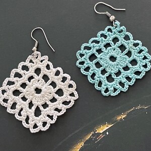 May include: A pair of crocheted earrings, one white and one light blue. The earrings are square shaped and have a delicate, intricate design.