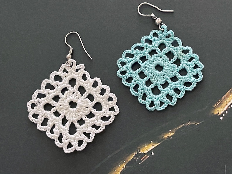 May include: A pair of crocheted earrings, one white and one light blue. The earrings are square shaped and have a delicate, intricate design.