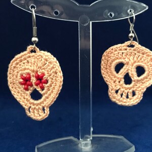 May include: A pair of crocheted skull earrings with red bead accents. The earrings are hanging from silver hooks on a clear display stand.
