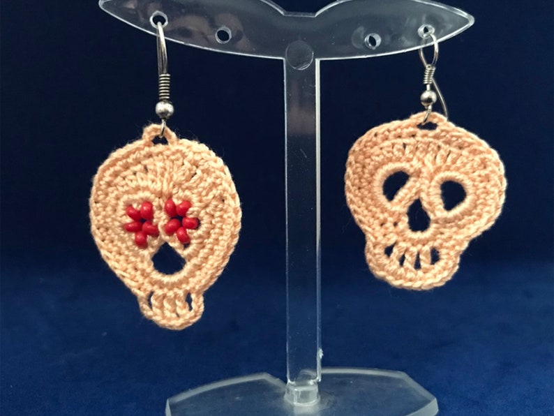 May include: A pair of crocheted skull earrings with red bead accents. The earrings are hanging from silver hooks on a clear display stand.