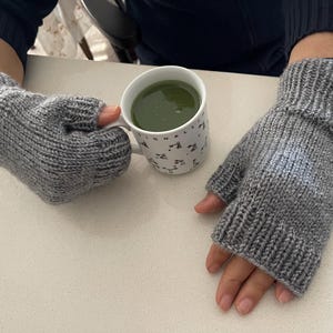 May include: A pair of gray knitted fingerless gloves. The gloves are made of a soft, textured yarn and have a ribbed cuff. A white mug with a green liquid and a black and white pattern is also visible.