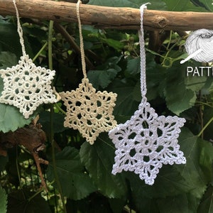 May include: Three crocheted snowflakes hanging from a branch. The snowflakes are white, cream, and beige. The snowflakes are hanging from a branch with green leaves.