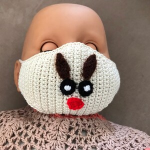 Puede incluir: Una m&aacute;scara facial de crochet blanca con un dise&ntilde;o de reno. La m&aacute;scara tiene cuernos marrones, ojos negros y una nariz roja.