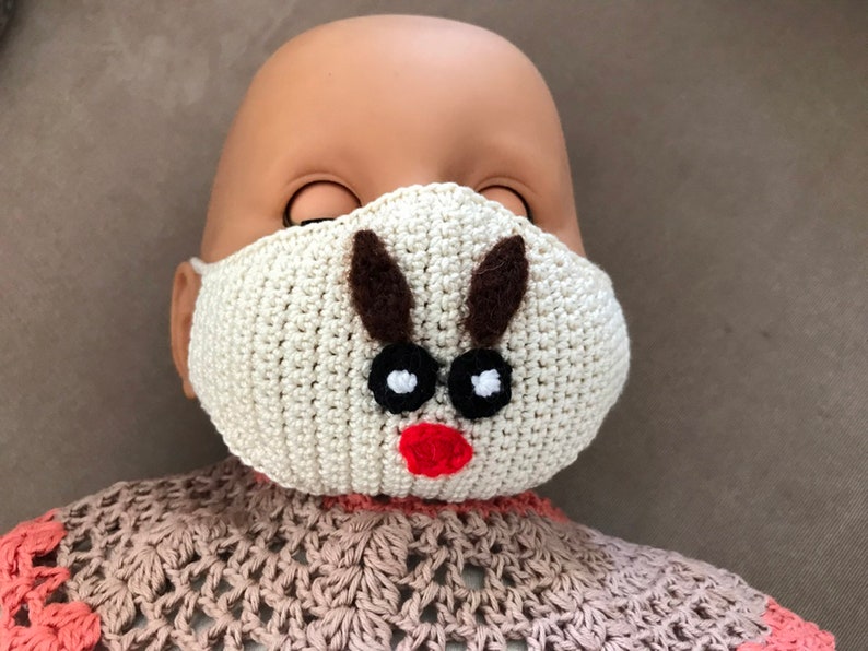 Puede incluir: Una m&aacute;scara facial de crochet blanca con un dise&ntilde;o de reno. La m&aacute;scara tiene cuernos marrones, ojos negros y una nariz roja.