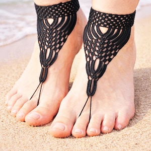 May include: Black crochet barefoot sandals. The sandals have an intricate woven design around the ankle and a delicate string that wraps around the big toe. The sandals are displayed on a sandy beach.