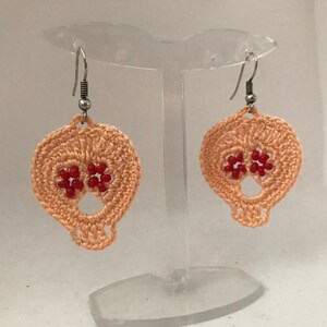 May include: A pair of peach-colored crocheted skull earrings with red bead accents. The earrings are hanging from silver hooks.