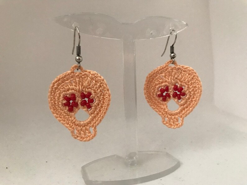 May include: A pair of peach-colored crocheted skull earrings with red bead accents. The earrings are hanging from silver hooks.