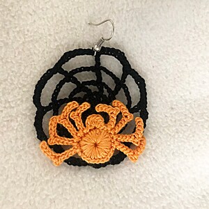 May include: A pair of crocheted earrings featuring a black web design with an orange spider in the center.