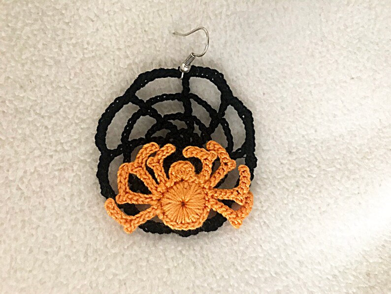 May include: A pair of crocheted earrings featuring a black web design with an orange spider in the center.