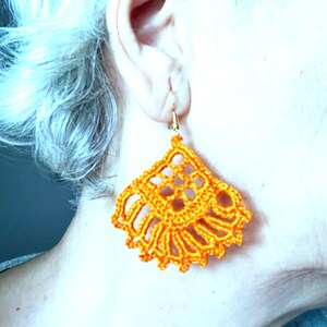 Puede incluir: Pendientes de ganchillo de color naranja con un dise&ntilde;o delicado e intrincado. Los pendientes cuelgan de la oreja de una persona.