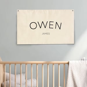 May include: A cream-colored banner with the name "OWEN" and the name "JAMES" printed in black. The banner is hanging above a wooden crib with a blanket and pillows. The room has a neutral color scheme with plants and a wooden shelf.