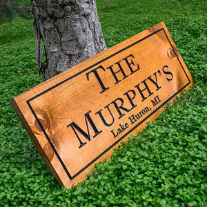 May include: A rectangular wooden sign with the text "THE MURPHY'S Lake Huron, MI" in black lettering. The sign is made of light brown wood with a dark brown border and is resting on a bed of green plants.