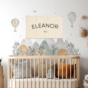 K&ouml;nnte beinhalten: Szene f&uuml;r die Wanddekoration im Kinderzimmer mit einem Holzbett und Kissen. Ein Banner mit den Namen "ELEANOR" und "MIA" h&auml;ngt &uuml;ber einem Berg-Wandbild. Hei&szlig;luftballon-Dekorationen und ein geflochtener Korb erg&auml;nzen die Szene.