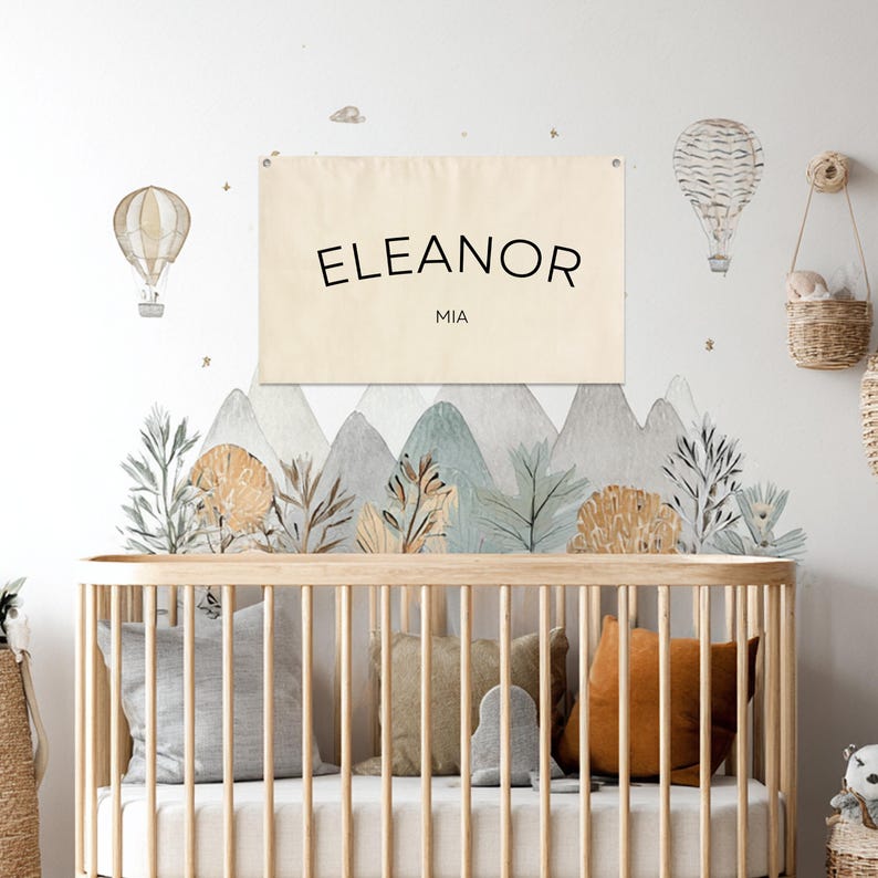 K&ouml;nnte beinhalten: Szene f&uuml;r die Wanddekoration im Kinderzimmer mit einem Holzbett und Kissen. Ein Banner mit den Namen "ELEANOR" und "MIA" h&auml;ngt &uuml;ber einem Berg-Wandbild. Hei&szlig;luftballon-Dekorationen und ein geflochtener Korb erg&auml;nzen die Szene.