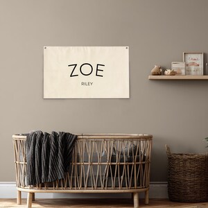 K&ouml;nnte beinhalten: Ein beiger Wandbehang aus Leinen mit dem Namen "ZOE" in schwarzen Buchstaben &uuml;ber dem Namen "RILEY". Der Wandbehang h&auml;ngt an einer neutralfarbenen Wand &uuml;ber einem Weidenkorb mit einer dunkelgrauen Decke.