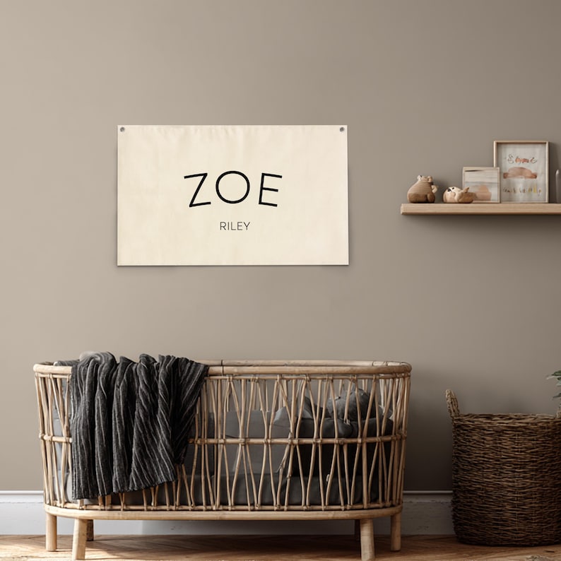 K&ouml;nnte beinhalten: Ein beiger Wandbehang aus Leinen mit dem Namen "ZOE" in schwarzen Buchstaben &uuml;ber dem Namen "RILEY". Der Wandbehang h&auml;ngt an einer neutralfarbenen Wand &uuml;ber einem Weidenkorb mit einer dunkelgrauen Decke.