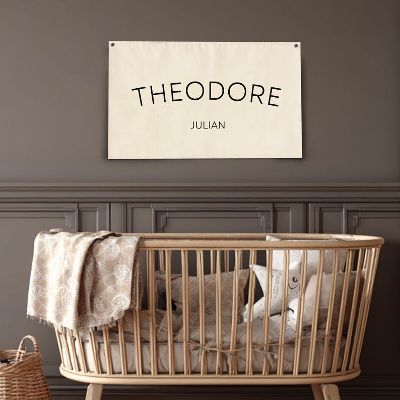 K&ouml;nnte beinhalten: Ein beiges Leinwandschild mit dem Namen "THEODORE" in Schwarz und dem Namen "JULIAN" darunter. Das Schild h&auml;ngt &uuml;ber einem Holzbettchen mit Bettw&auml;sche und Kissen. Ein Weidenkorb steht auf dem Boden.
