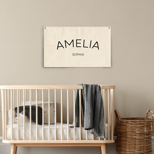 K&ouml;nnte beinhalten: Ein cremefarbenes Banner mit dem Namen "AMELIA" in Schwarz und "SOPHIA" darunter. Das Banner h&auml;ngt &uuml;ber einem Holzbettchen mit Bettw&auml;sche, einem Kissen und einer Decke. Rechts befindet sich ein Weidenkorb.