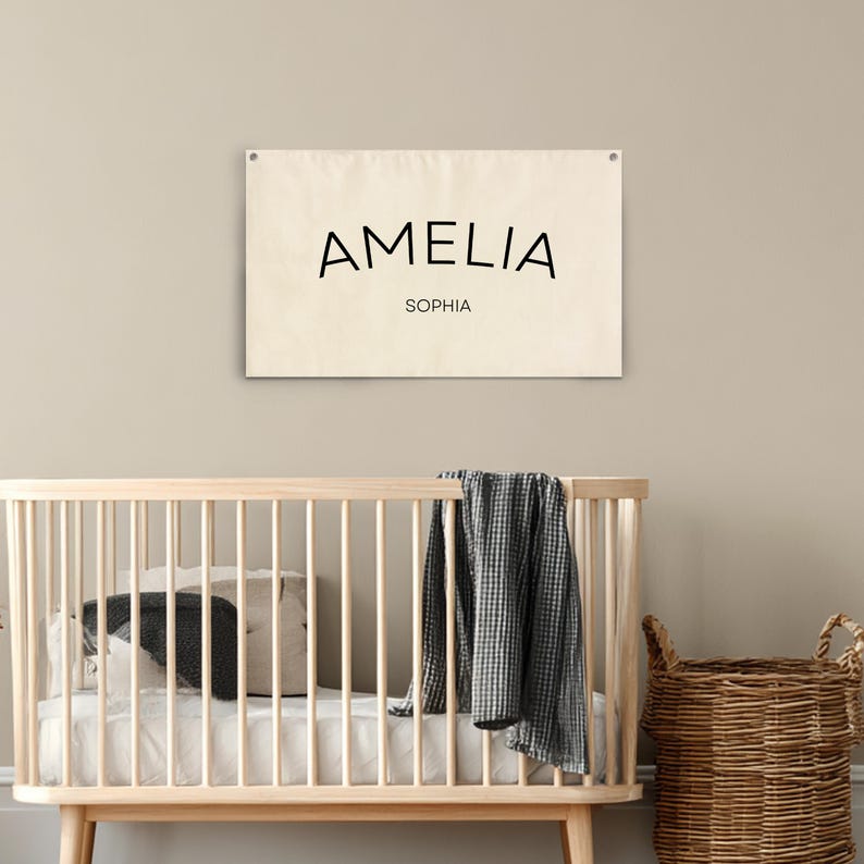 K&ouml;nnte beinhalten: Ein cremefarbenes Banner mit dem Namen "AMELIA" in Schwarz und "SOPHIA" darunter. Das Banner h&auml;ngt &uuml;ber einem Holzbettchen mit Bettw&auml;sche, einem Kissen und einer Decke. Rechts befindet sich ein Weidenkorb.