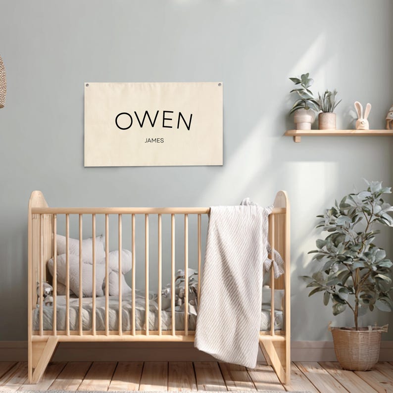 K&ouml;nnte beinhalten: Ein cremefarbenes Banner mit dem Namen "OWEN" und dem Namen "JAMES" in schwarzer Schrift. Das Banner h&auml;ngt &uuml;ber einem Holzbettchen mit einer Decke und Kissen. Der Raum hat ein neutrales Farbschema mit Pflanzen und einem Holzregal.
