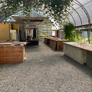 May include: Interior view of a greenhouse with a gravel floor and wooden tables. The structure has a curved, translucent roof and is adorned with hanging vines. Sunlight streams through, illuminating the space. A colorful sign is visible in the background.