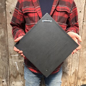 May include: A black square slate sign with a metal hanging hook on the back. The sign is being held by a person wearing a red and black plaid shirt and blue jeans.