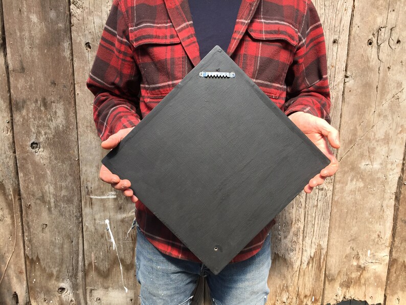 May include: A black square slate sign with a metal hanging hook on the back. The sign is being held by a person wearing a red and black plaid shirt and blue jeans.