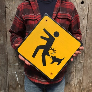 May include: A yellow diamond-shaped warning sign with a black silhouette of a person being kicked in the rear end by a dog. The sign has a black border and is held by a person wearing a red and black plaid shirt.