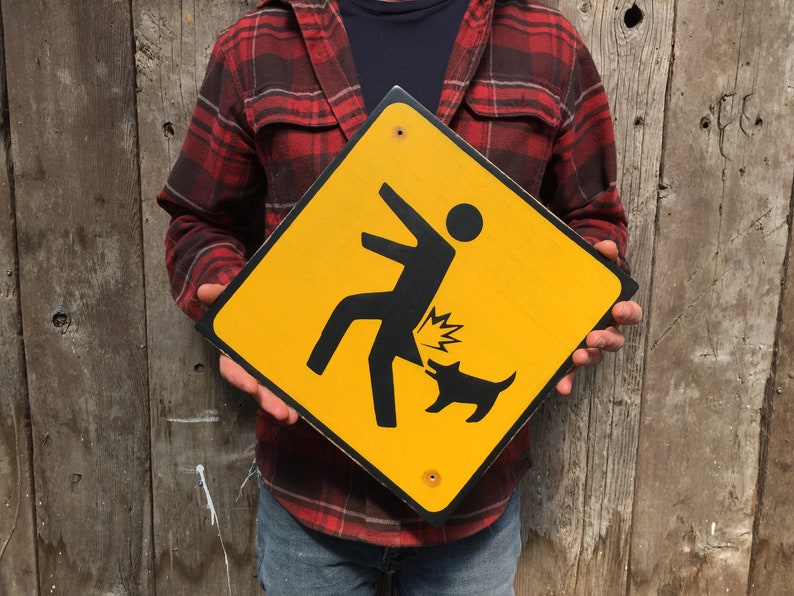 May include: A yellow diamond-shaped warning sign with a black silhouette of a person being kicked in the rear end by a dog. The sign has a black border and is held by a person wearing a red and black plaid shirt.