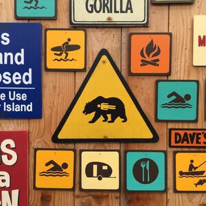 May include: A collection of colorful wooden signs with various icons and text. Signs include a bear warning, a surfer, a swimmer, a campfire, a cabin, and text that reads "This Island Closed" and "This is a Sign."