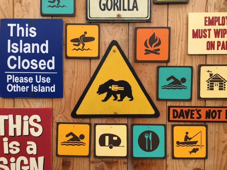 May include: A collection of colorful wooden signs with various icons and text. Signs include a bear warning, a surfer, a swimmer, a campfire, a cabin, and text that reads "This Island Closed" and "This is a Sign."