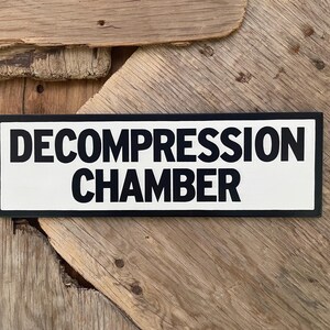 May include: A rectangular sign with the words "DECOMPRESSION CHAMBER" in bold, black capital letters on a white background. The sign has a black border and is set against a wooden background.