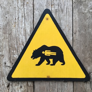 May include: A yellow triangular warning sign with a black border. The sign features a black silhouette of a bear with a human figure inside. The sign is mounted on a weathered wooden surface.