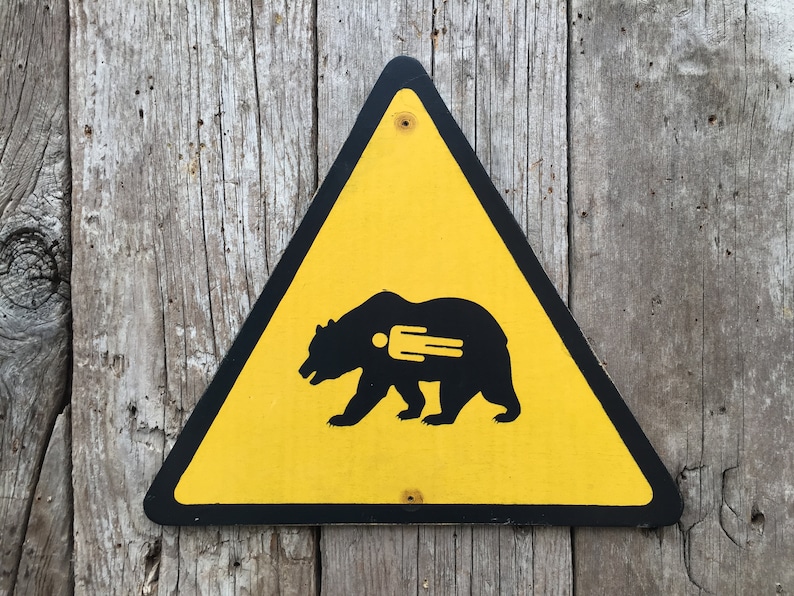 May include: A yellow triangular warning sign with a black border. The sign features a black silhouette of a bear with a human figure inside. The sign is mounted on a weathered wooden surface.