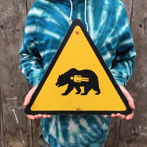 May include: A yellow triangular sign with a black border and a black bear silhouette with a fork inside. The sign is held in front of a wooden background. The bear sign is a warning sign.