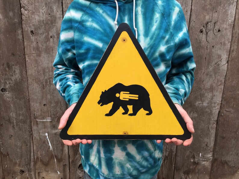 May include: A yellow triangular sign with a black border and a black bear silhouette with a fork inside. The sign is held in front of a wooden background. The bear sign is a warning sign.