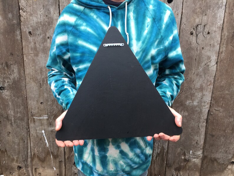 May include: A black, triangular wooden sign with a sawtooth hanger on the back. The sign is held up against a wooden background. The sign is a solid black color.