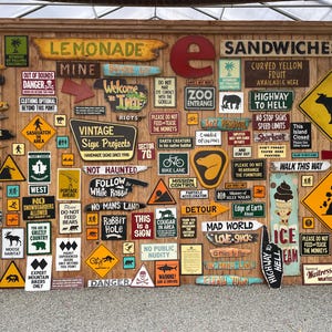 May include: A collection of vintage-style wooden signs with various messages and graphics, including "Lemonade," "Sandwiches," and directional arrows. The signs are mounted on a wooden wall, creating a rustic and eclectic display.