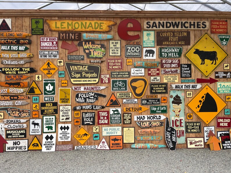 May include: A collection of vintage-style wooden signs with various messages and graphics, including "Lemonade," "Sandwiches," and directional arrows. The signs are mounted on a wooden wall, creating a rustic and eclectic display.