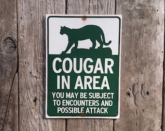 May include: A green and white warning sign with a silhouette of a cougar. The sign reads "Cougar in Area. You may be subject to encounters and possible attack."