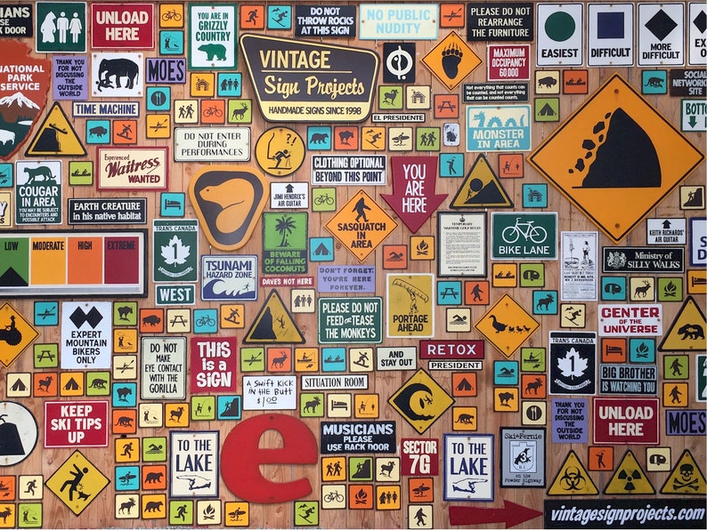 May include: A collection of vintage signs on a wooden background. The signs feature various warnings, directions, and humorous messages. The signs include text such as "Unload Here", "Grizzly Country", and "Bike Lane". The signs are in various shapes and colors.