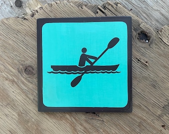 May include: Square wooden sign with a black border and a turquoise background. The sign features a black silhouette of a person kayaking, with a paddle in the water. The sign is placed on a weathered wooden surface.
