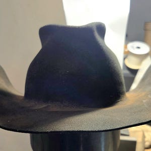 May include: A dark charcoal gray cowboy hat with a high, pinched crown and a wide, curved brim. The hat features a matte, textured finish with subtle lighter brown weathering along the edges of the brim and the base of the crown.