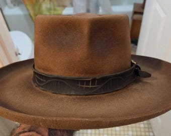 May include: A brown felt fedora hat with a wide brim and a pinched crown. The hat features a dark brown leather band wrapped around the base, decorated with a repeating etched wave and vertical line pattern and a small side buckle closure.