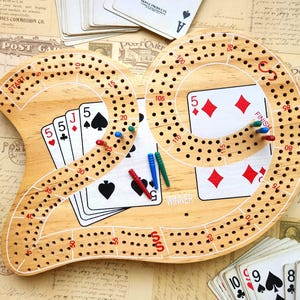 May include: A light-colored wooden cribbage board shaped like the number 29, featuring red printed numbers, the words START, FINISH, and WINNER, and multiple rows of holes. Several colored plastic pegs are inserted into the board surface.