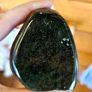 May include: A dark green, polished, smooth, oval-shaped stone with a slight sheen. The stone has a few white spots and a light line running through it.
