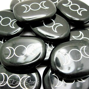 以下が含まれることがあります： 黒く磨かれた石の集まり。それぞれに白いトリプルムーンのシンボルが刻まれています。石は滑らかで楕円形です。