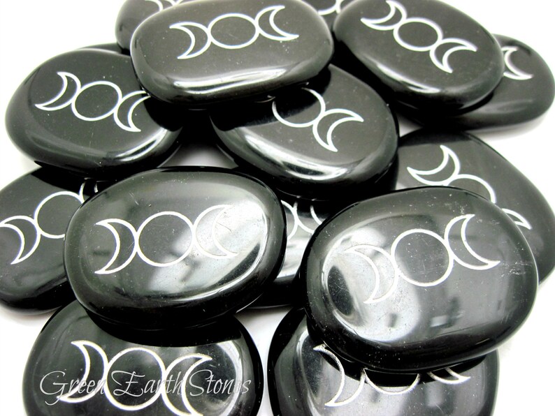 以下が含まれることがあります： 黒く磨かれた石の集まり。それぞれに白いトリプルムーンのシンボルが刻まれています。石は滑らかで楕円形です。